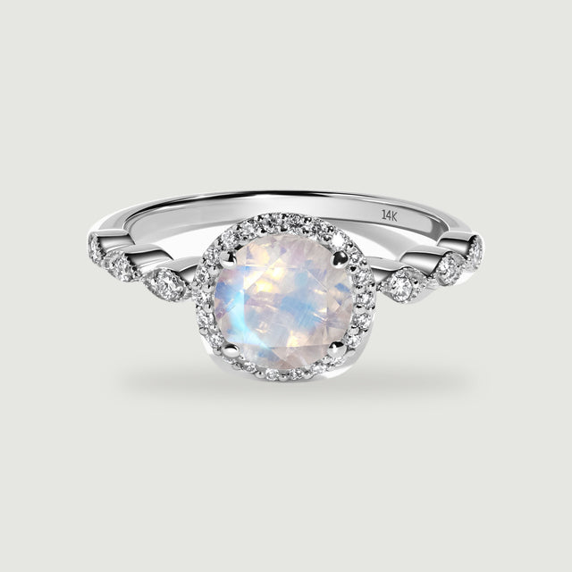 A white gold ring with a round faceted moonstone, a diamond halo and scalloped diamond shoulders rests on light gray.