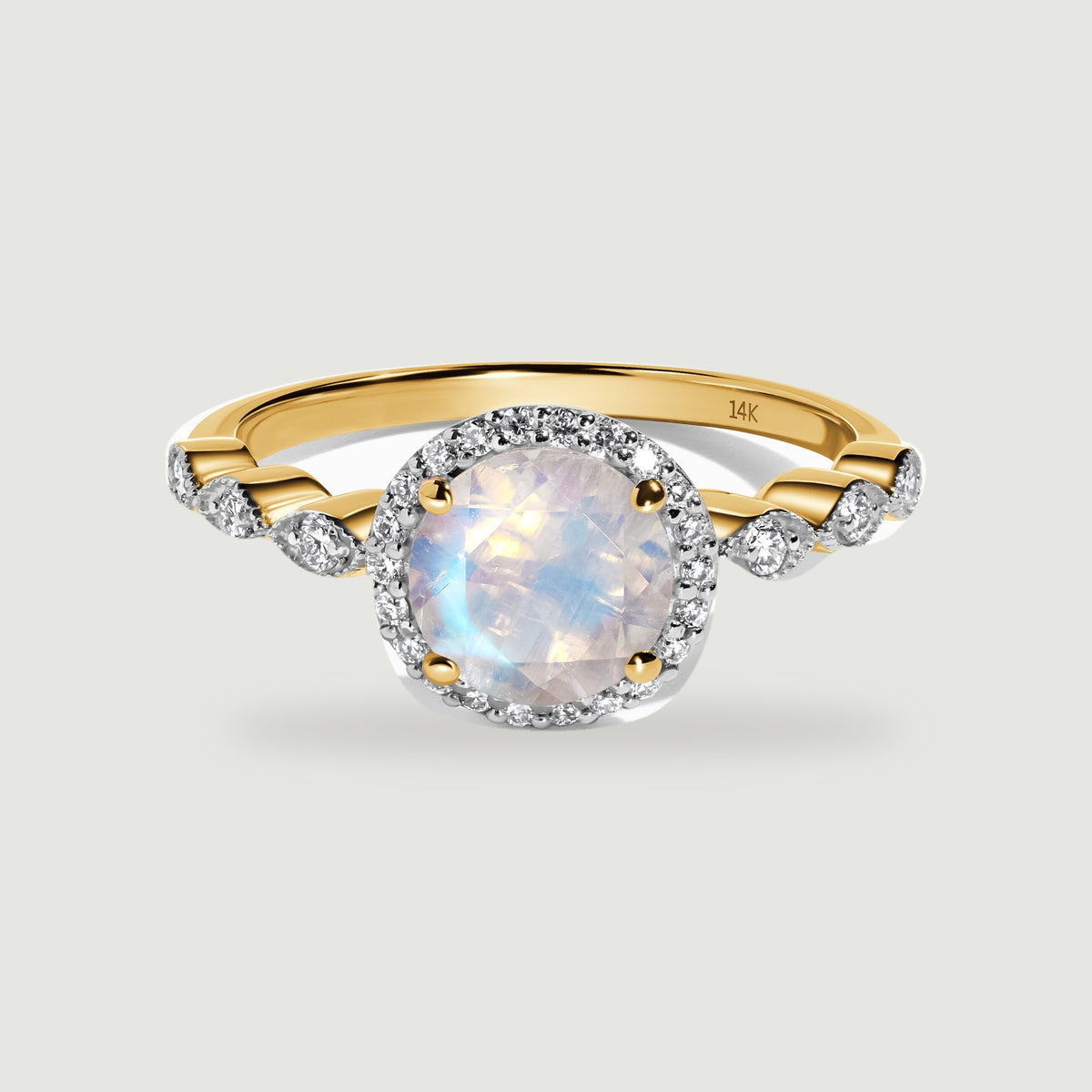 A yellow gold ring with a pale rainbow moonstone and diamond halo is centered on a light gray surface.