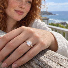 A rose gold moonstone ring with a diamond halo is shown on a freckled hand resting on weathered wood by the sea.