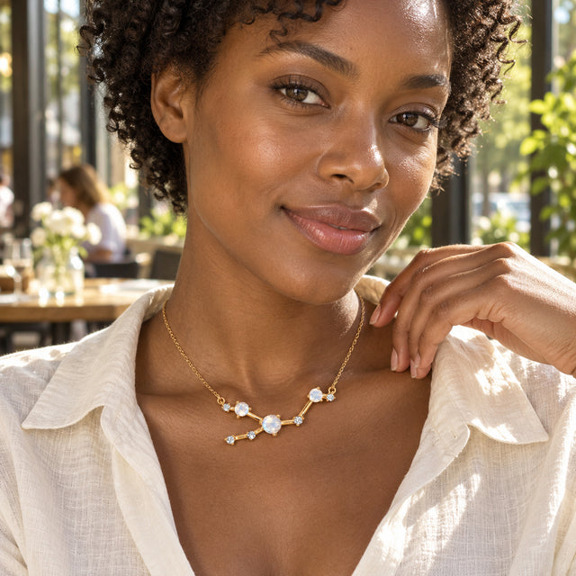 A woman with short curls wears the yellow gold moonstone necklace with a white linen shirt in a sunlit cafe.