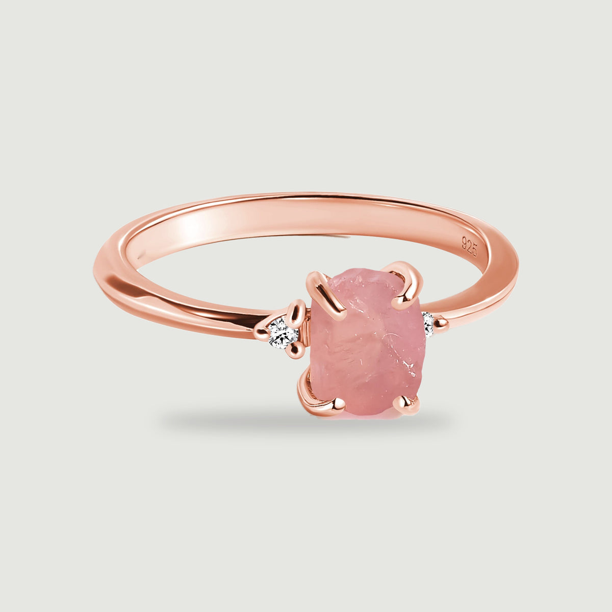A complete rose gold ring has a rough pink center stone and two round clear accents on a light gray surface.