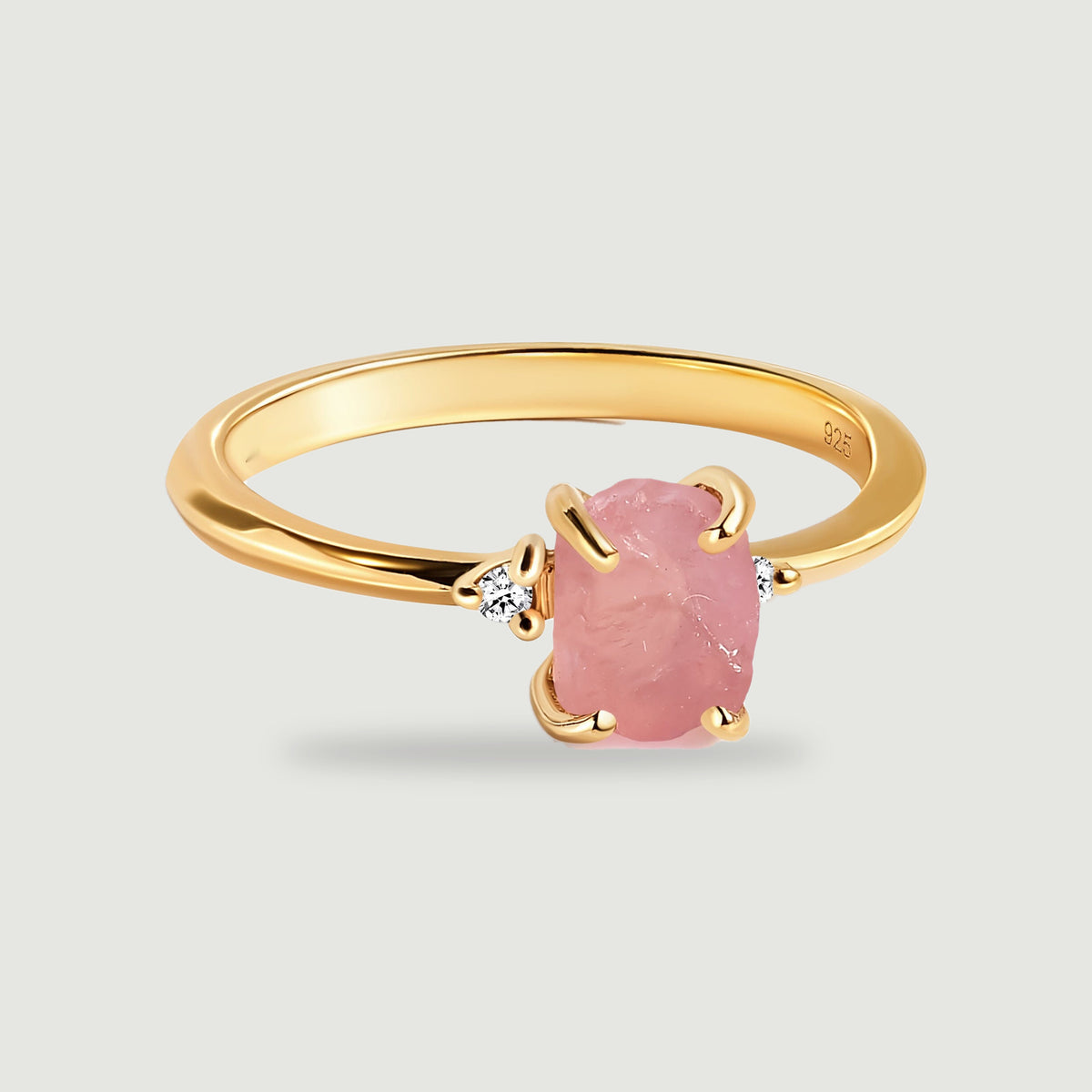 A complete yellow gold ring has a rough pink center stone and two round clear accents on a light gray surface.