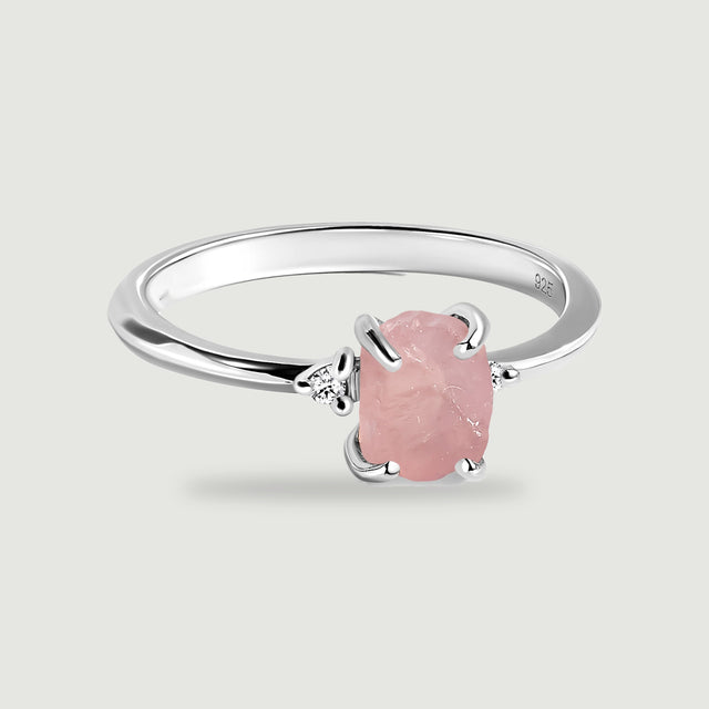 A complete silver ring has a rough pink center stone and two round clear accents on a light gray surface.