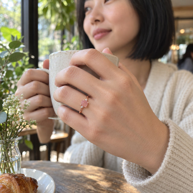 A rose gold ring with a pink center stone is worn on a hand holding a ceramic cup beside a cafe window.