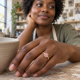 A yellow gold ring with a pink stone and two clear accents is shown on a hand resting beside a clay bowl.