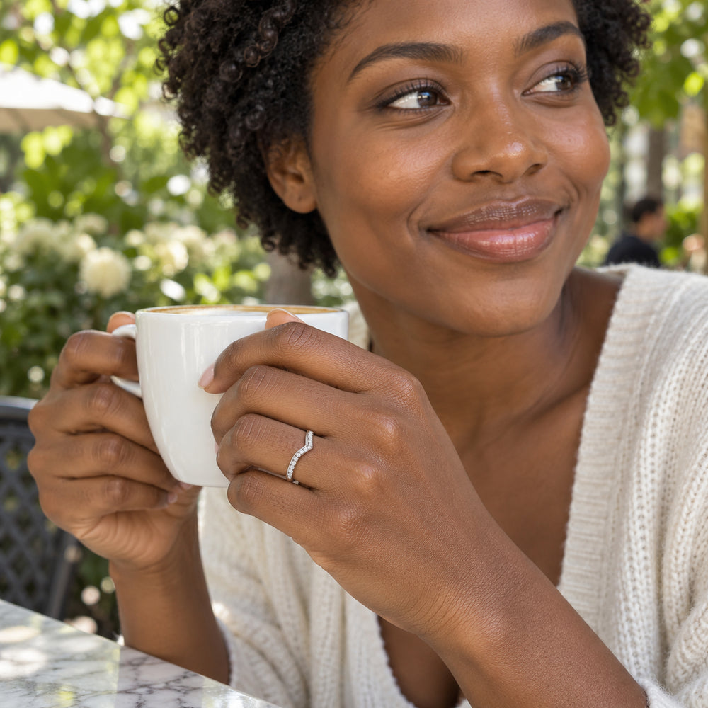 A white gold chevron diamond ring is worn on a hand holding a white coffee cup at a leafy cafe.