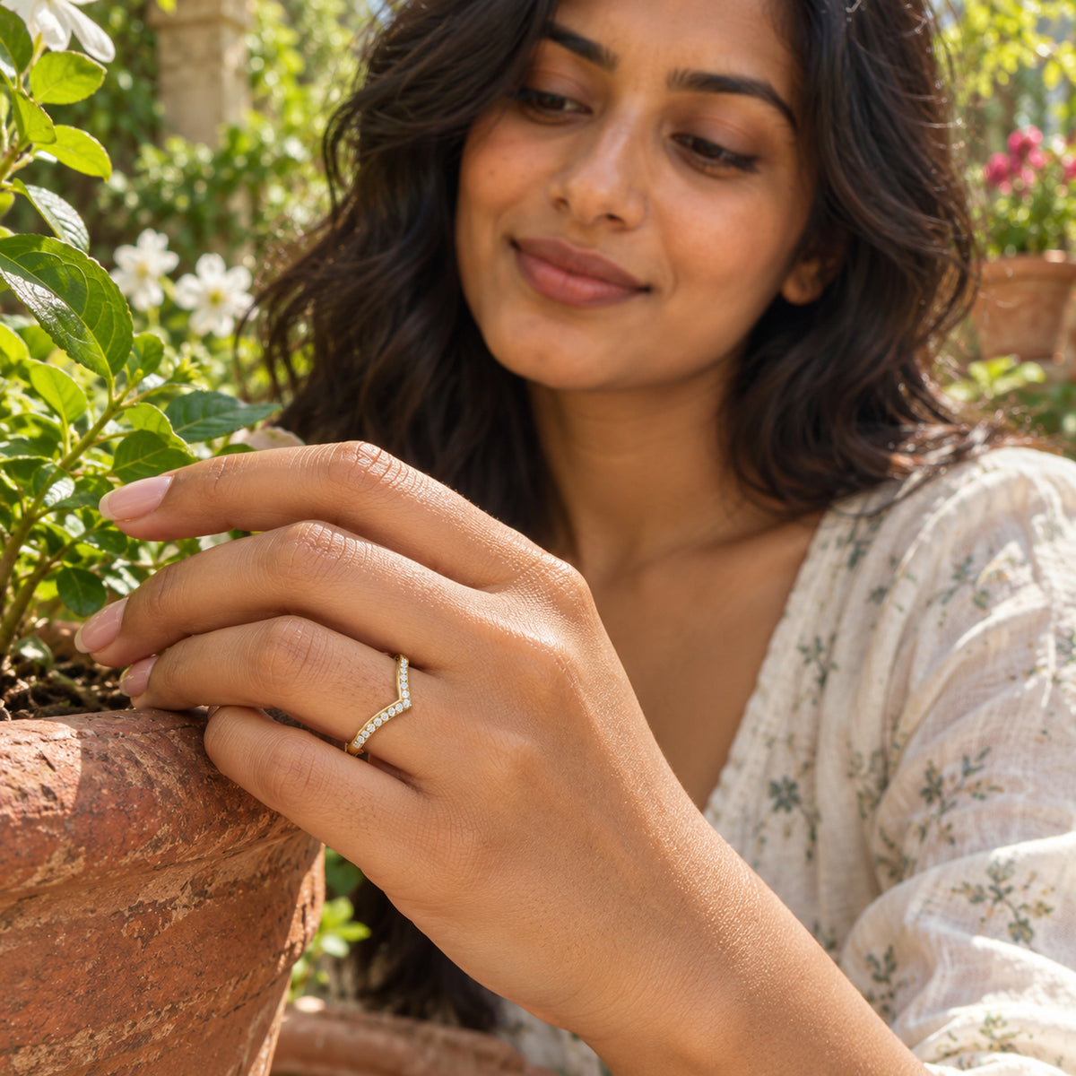 A yellow gold diamond chevron ring is shown on a hand resting against a terracotta pot in a sunlit garden.