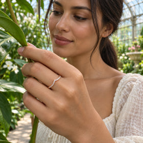 A rose gold diamond chevron ring is worn on a hand holding a leafy stem inside a glass conservatory.