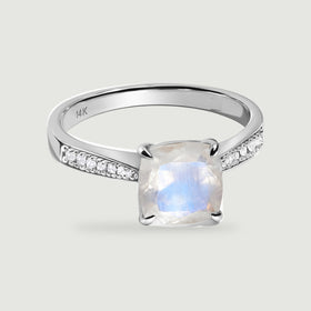 A white gold ring with a cushion-shaped moonstone and diamond-set shoulders rests on a pale gray surface.