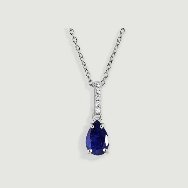 A silver link chain holds a blue pear-shaped pendant beneath a vertical row of five clear stones on a pale gray background.