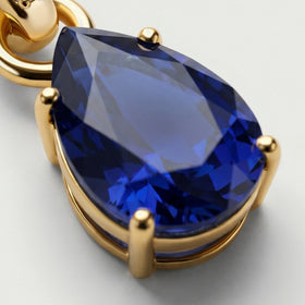 An angled detail reveals the raised gold prongs and the rim beneath the deep blue pear-shaped sapphire.