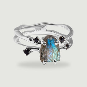 A sterling silver ring holds a raw blue-green labradorite between four black accent stones on a light gray background.