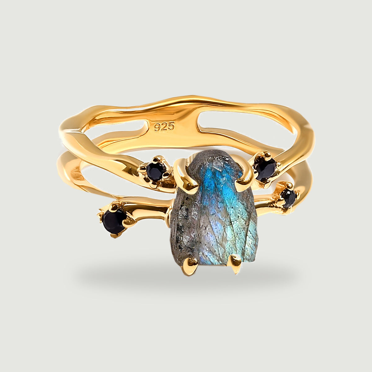 A yellow gold vermeil ring holds a raw blue-green labradorite between four black accent stones on a light gray background.