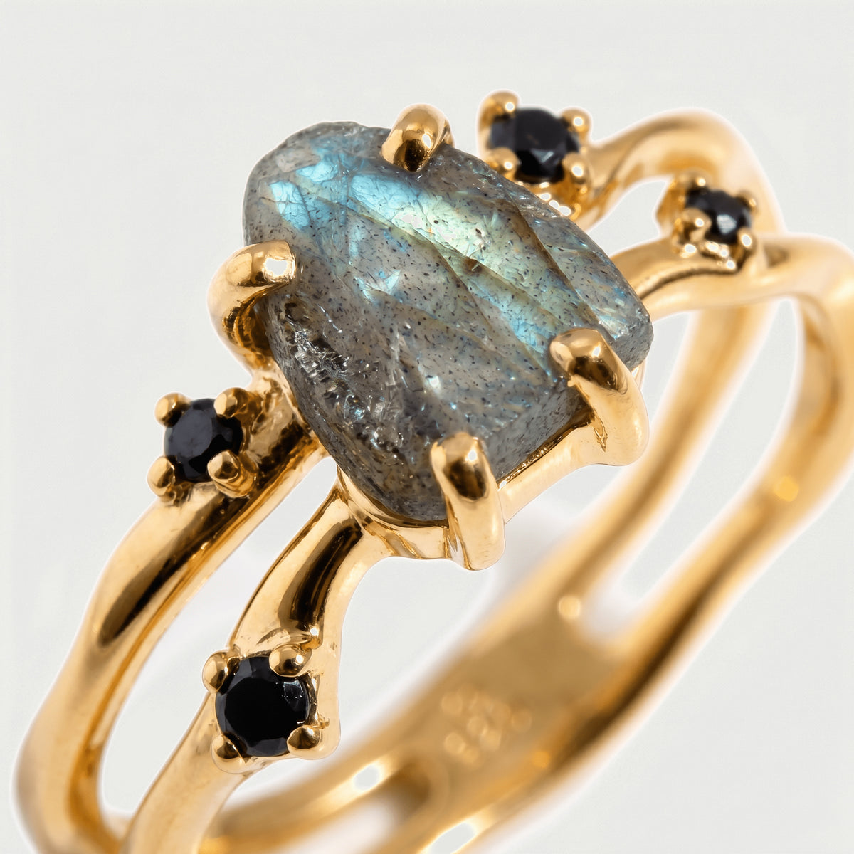 Raw labradorite with blue and green flashes sits in rounded yellow gold prongs beside small black stones.