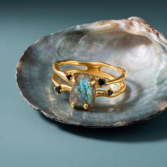 The yellow gold labradorite ring is arranged on a pearly shell over a clean blue tabletop.