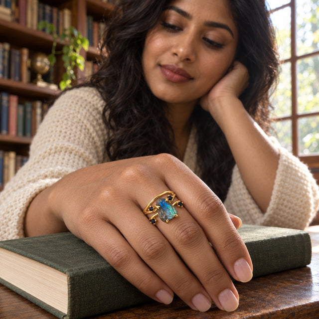 A yellow gold labradorite ring is shown on a woman's hand resting on a closed green book beside library shelves.
