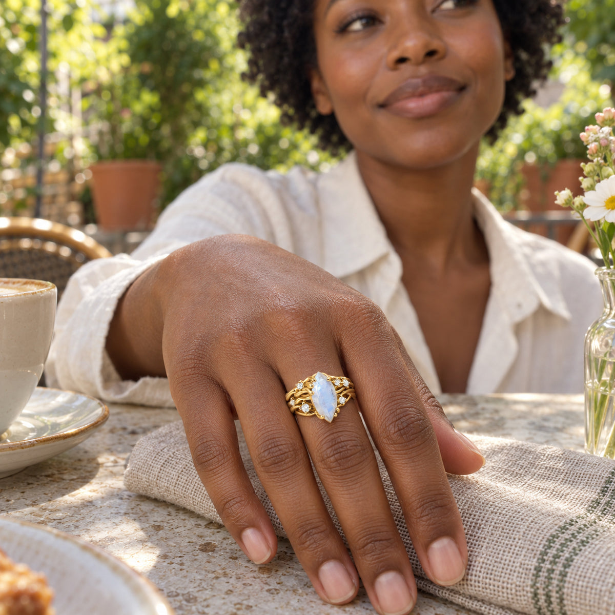 A yellow gold vermeil moonstone ring is worn on a hand resting on a linen napkin at an outdoor cafe.