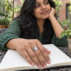 A silver moonstone ring is shown on a hand resting on an open sketchbook in a leafy brick courtyard.