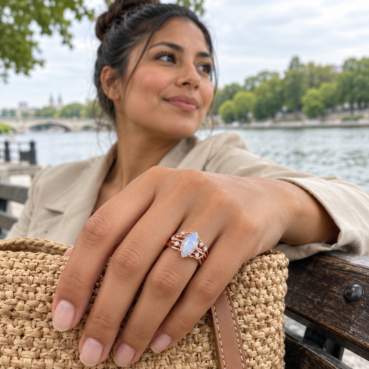 A rose gold vermeil ring with a pale blue moonstone is worn beside a woven bag on a riverside bench.