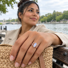 A rose gold vermeil ring with a pale blue moonstone is worn beside a woven bag on a riverside bench.