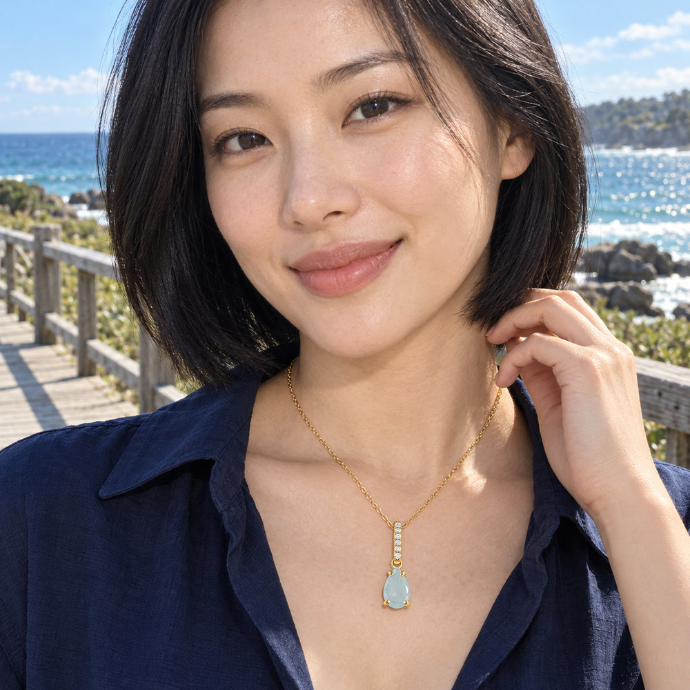 A woman in a navy shirt wears the yellow gold aquamarine necklace beside a seaside boardwalk.