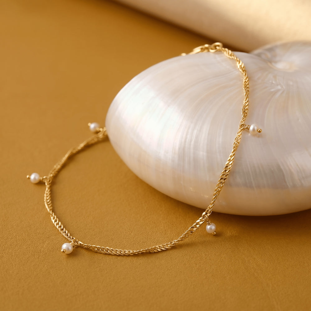 A gold pearl anklet is draped over a white shell on a clean warm toned surface.