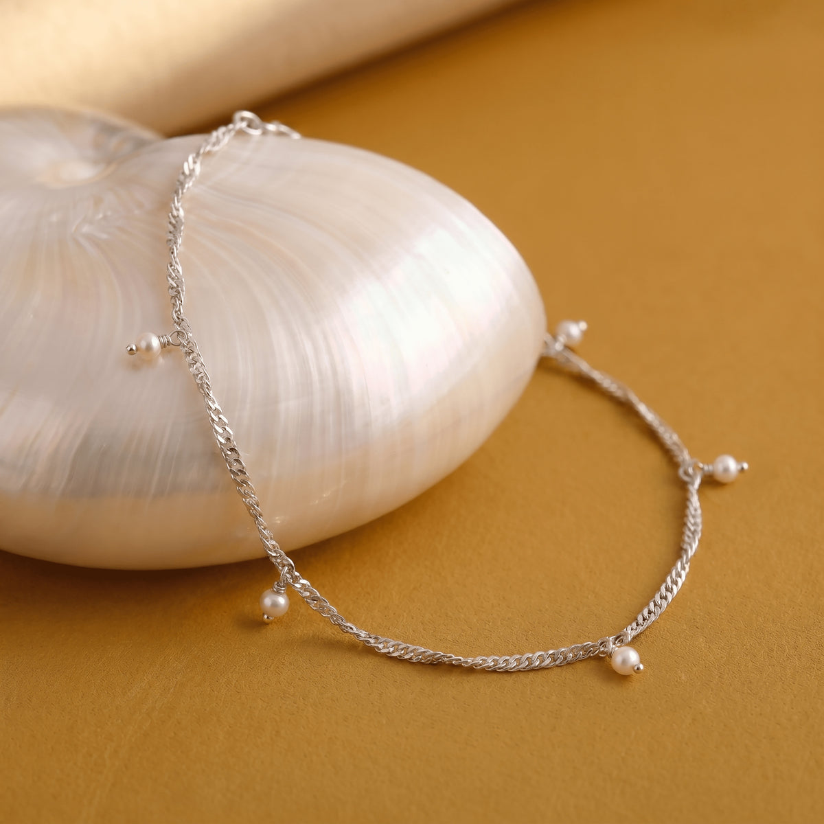 A silver chain with small pearl charms curves around a white shell on a warm tabletop.