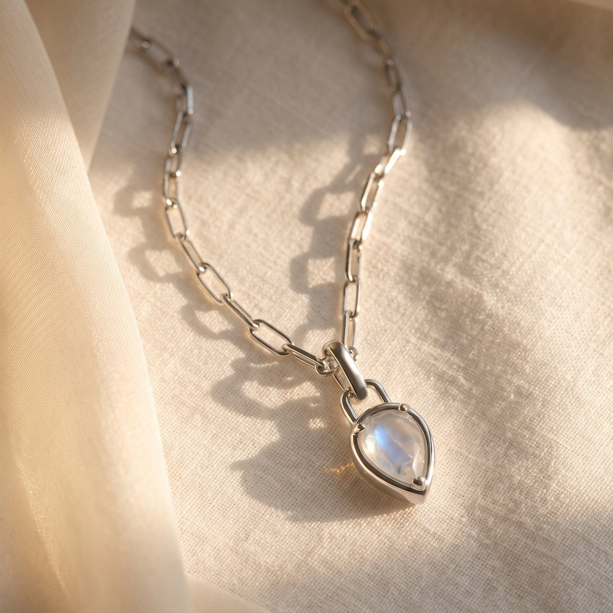 The silver-tone moonstone necklace rests on cream woven fabric beside a soft ribbon in natural light.