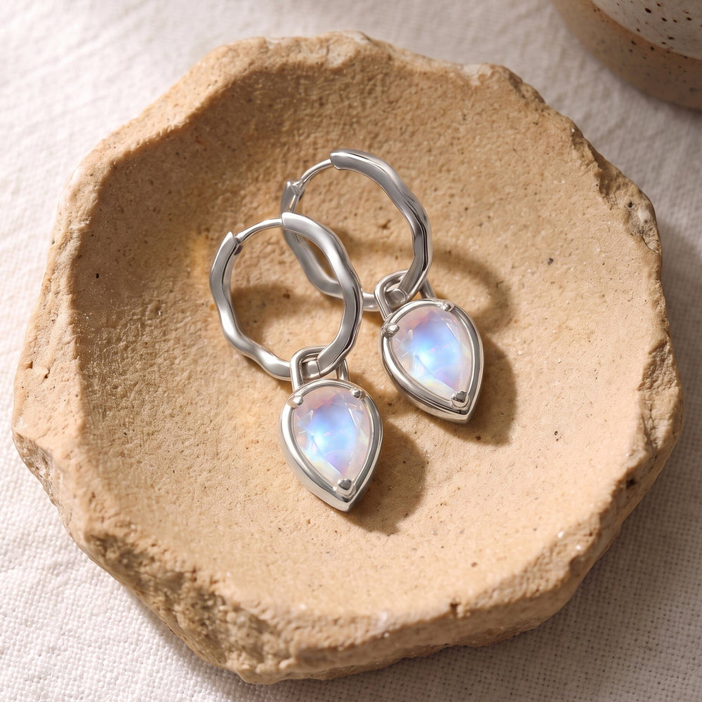 Two silver-tone moonstone hoop earrings rest together in a textured beige ceramic dish.