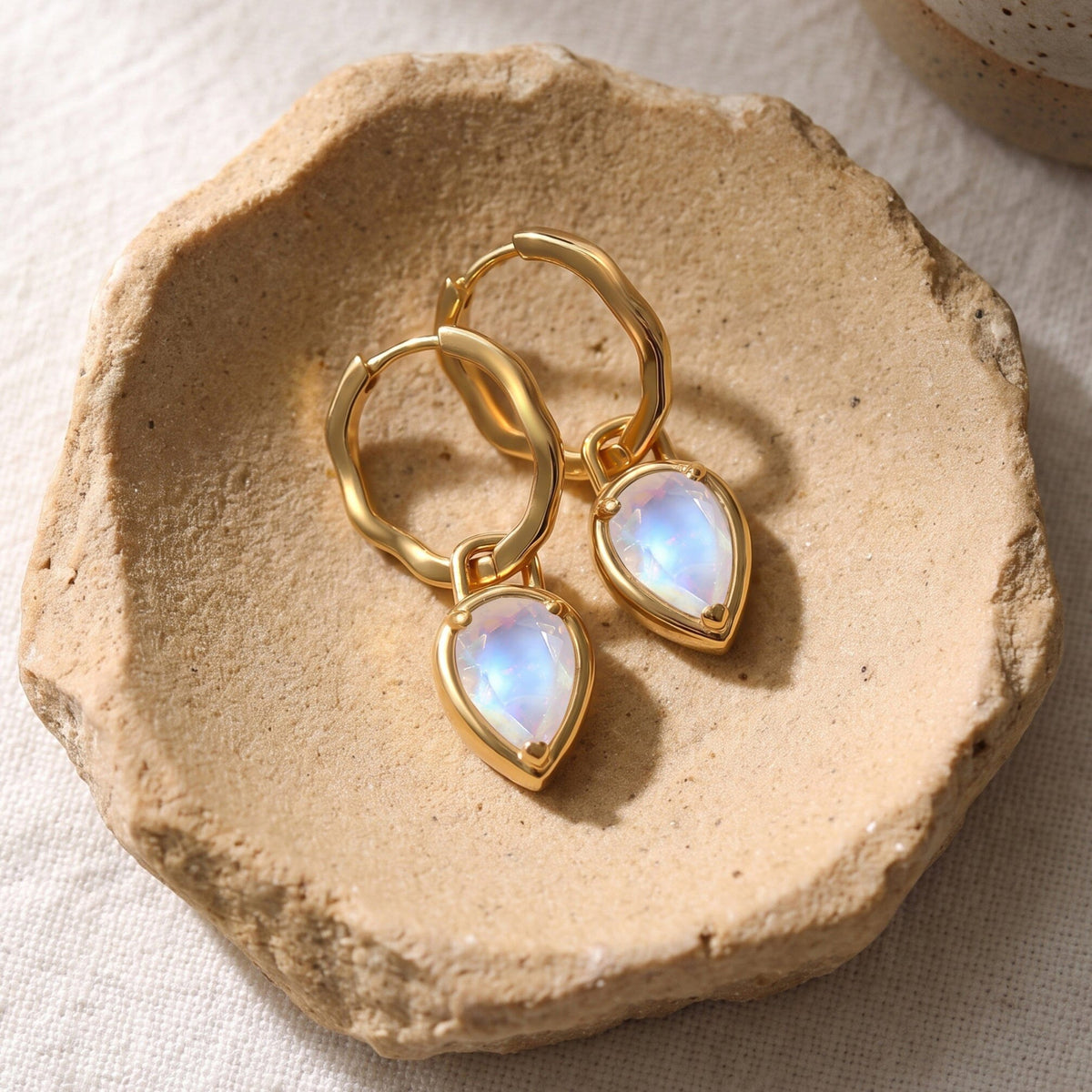 Yellow-gold-tone moonstone earrings lie side by side on a sand-coloured ceramic dish.