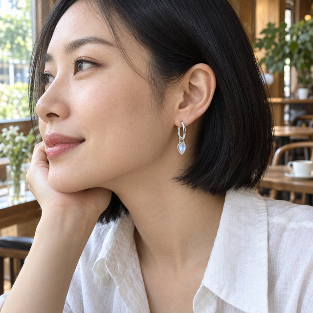 A woman with a short dark bob wears a silver-tone hoop with a pear-shaped moonstone drop beside a cafe window.