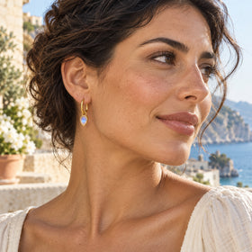 A woman with swept-back wavy hair wears a yellow-gold-tone moonstone hoop on a terrace overlooking the sea.