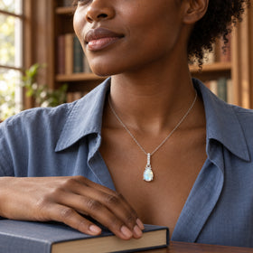 A woman in a blue linen shirt wears the silver moonstone pendant beside a window, with one hand resting on a closed book.