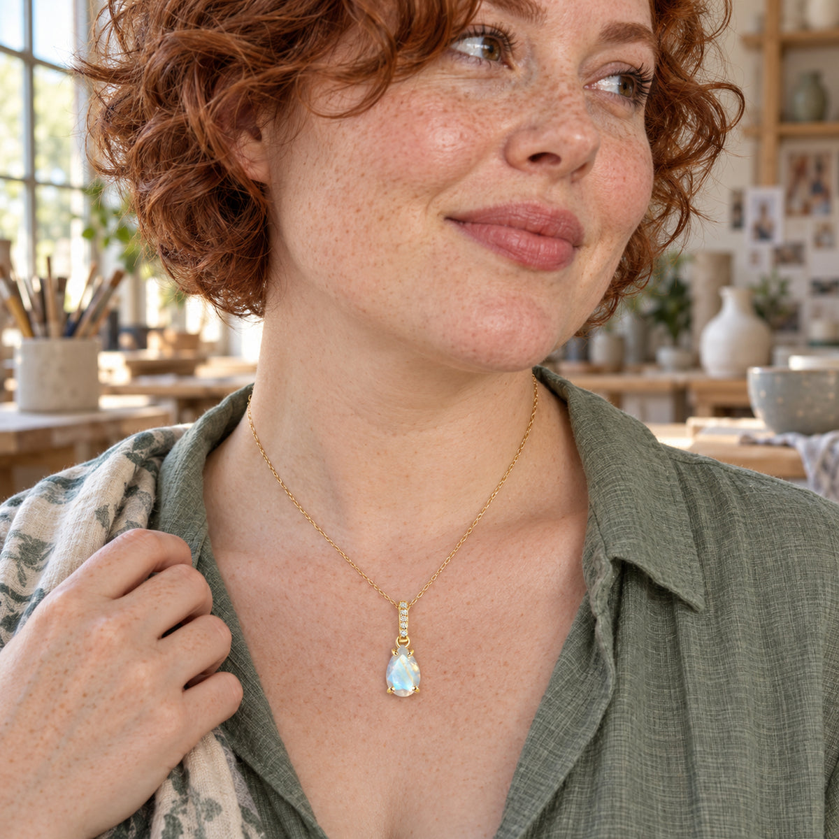 A woman with short red curls wears the yellow gold vermeil moonstone necklace over a green shirt in a sunlit ceramics studio.