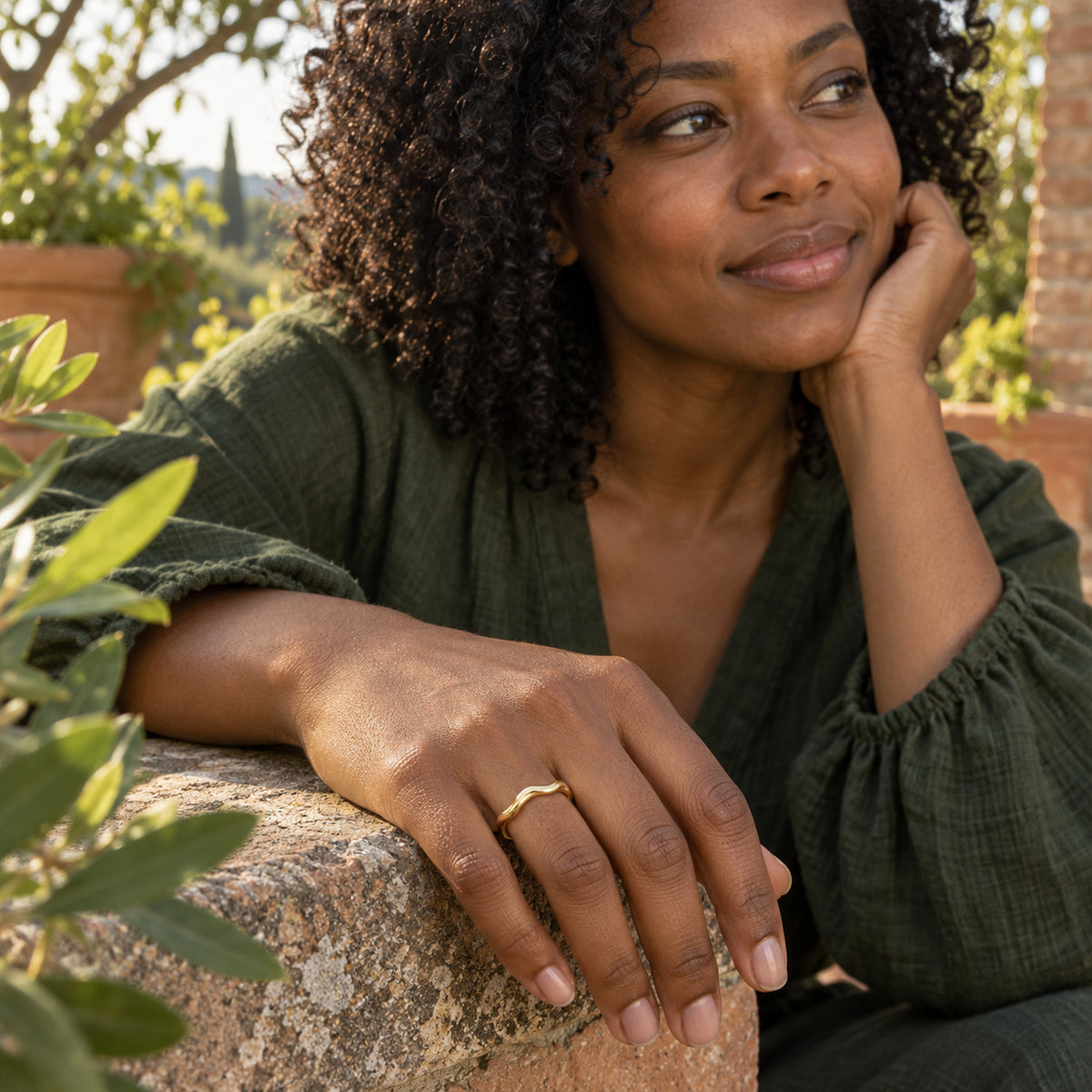 A polished yellow gold wavy band is worn on a hand resting on a stone ledge beside green leaves.