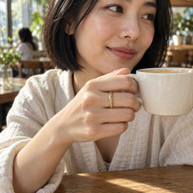 A yellow gold wave band is visible on a finger holding an ivory ceramic cup in a sunlit cafe.