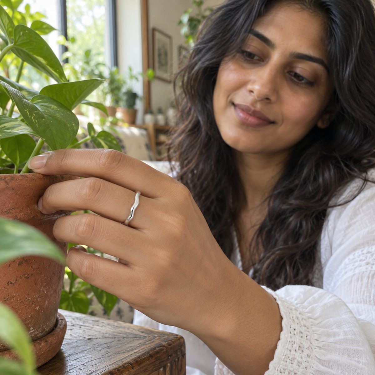 A polished silver wavy ring is worn on a hand touching a terracotta plant pot near a window.