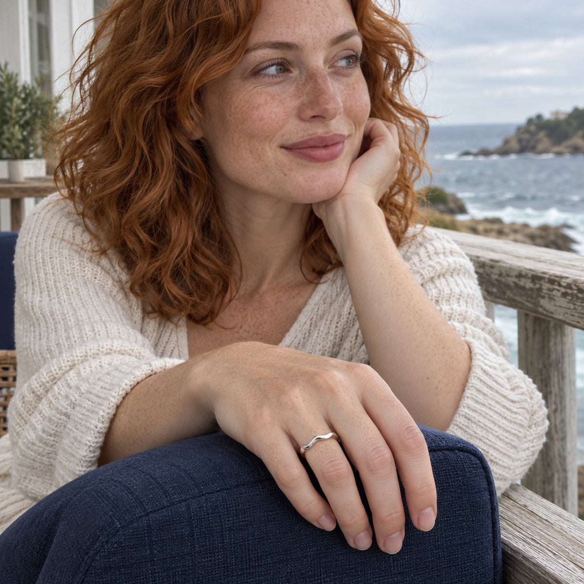A silver wave band is visible on a relaxed hand resting on navy linen beside a seaside balcony rail.