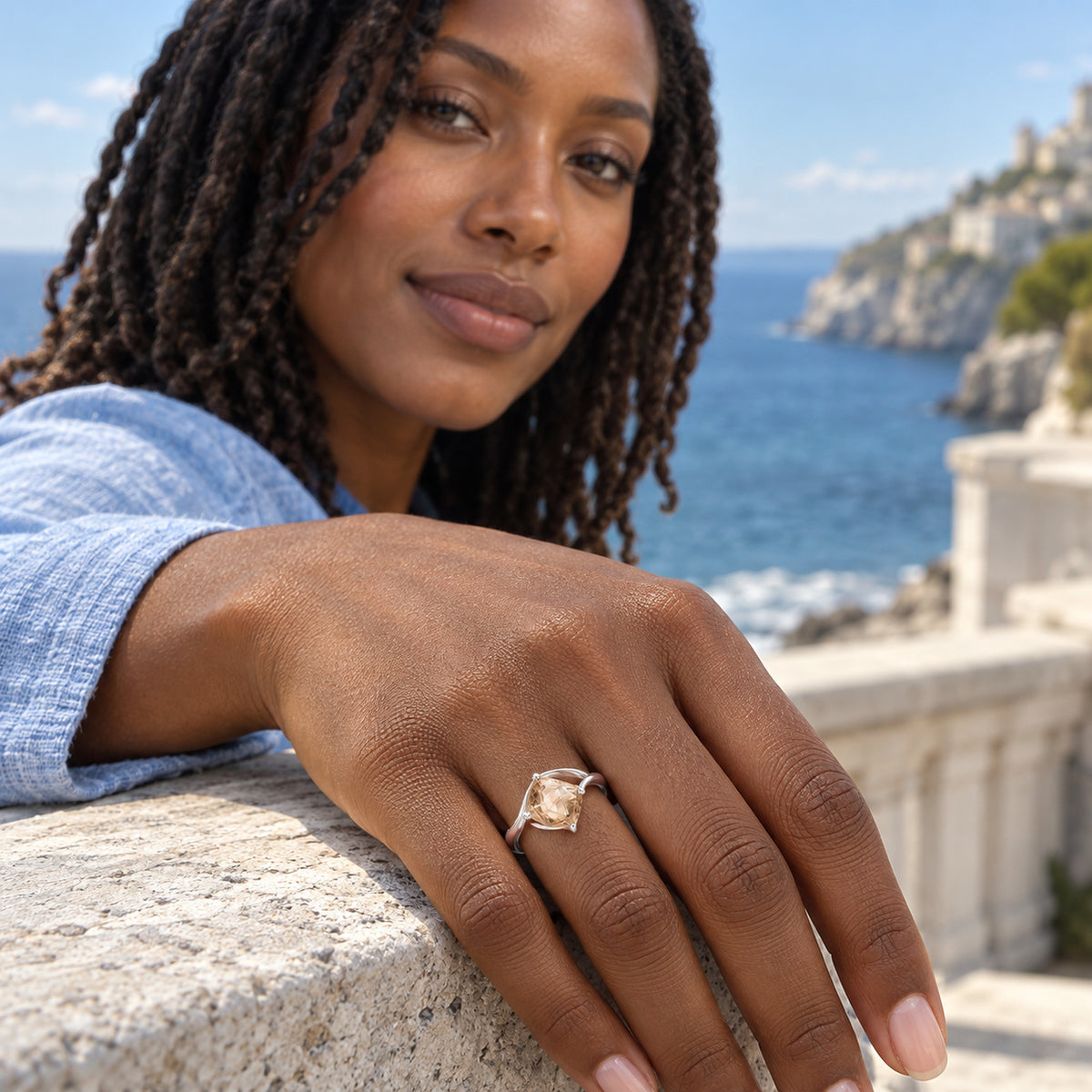 A woman with twists wears the sterling silver champagne quartz ring while resting her hand on a stone railing above the sea.