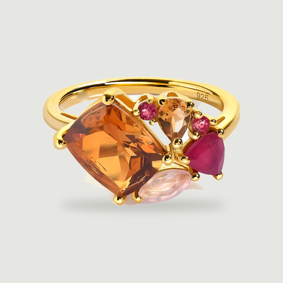 A yellow gold ring displays a large orange rectangular gemstone beside smaller pink and amber stones on a light gray surface.