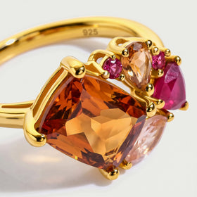 A close view shows orange and pink gemstone facets held by rounded yellow gold prongs.