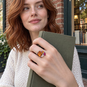 A red haired woman wears the silver orange and pink gemstone ring while holding a plain clothbound book outside a brick storefront.