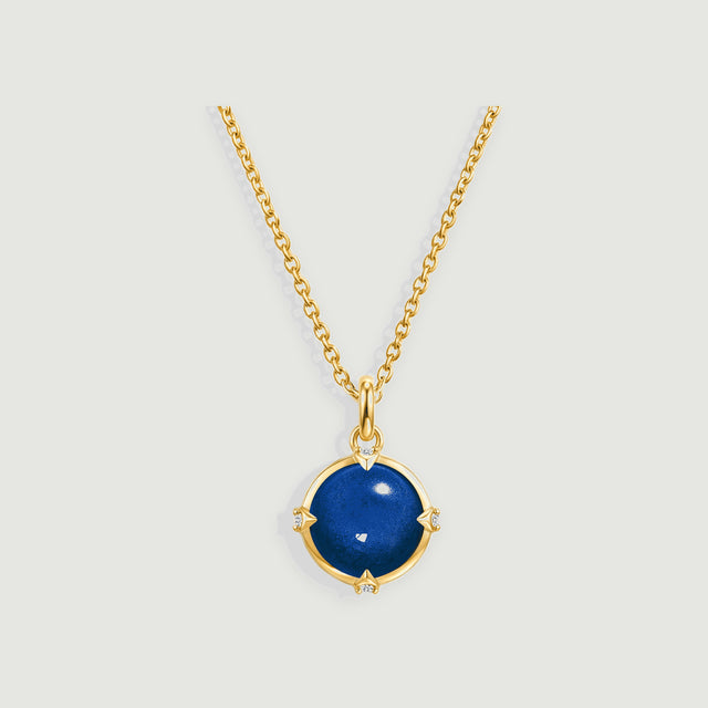 A round blue pendant in a yellow gold setting hangs from a fine chain on a light gray background.