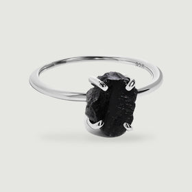 A raw black obsidian stone rests in four silver prongs on a slender polished ring against a light gray background.