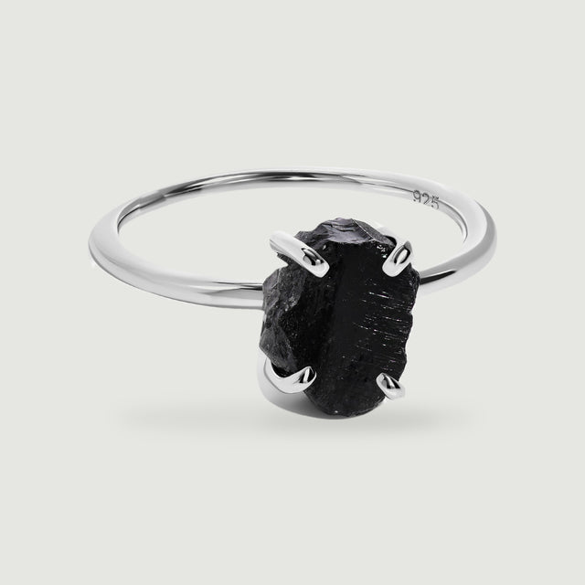 A raw black obsidian stone rests in four silver prongs on a slender polished ring against a light gray background.