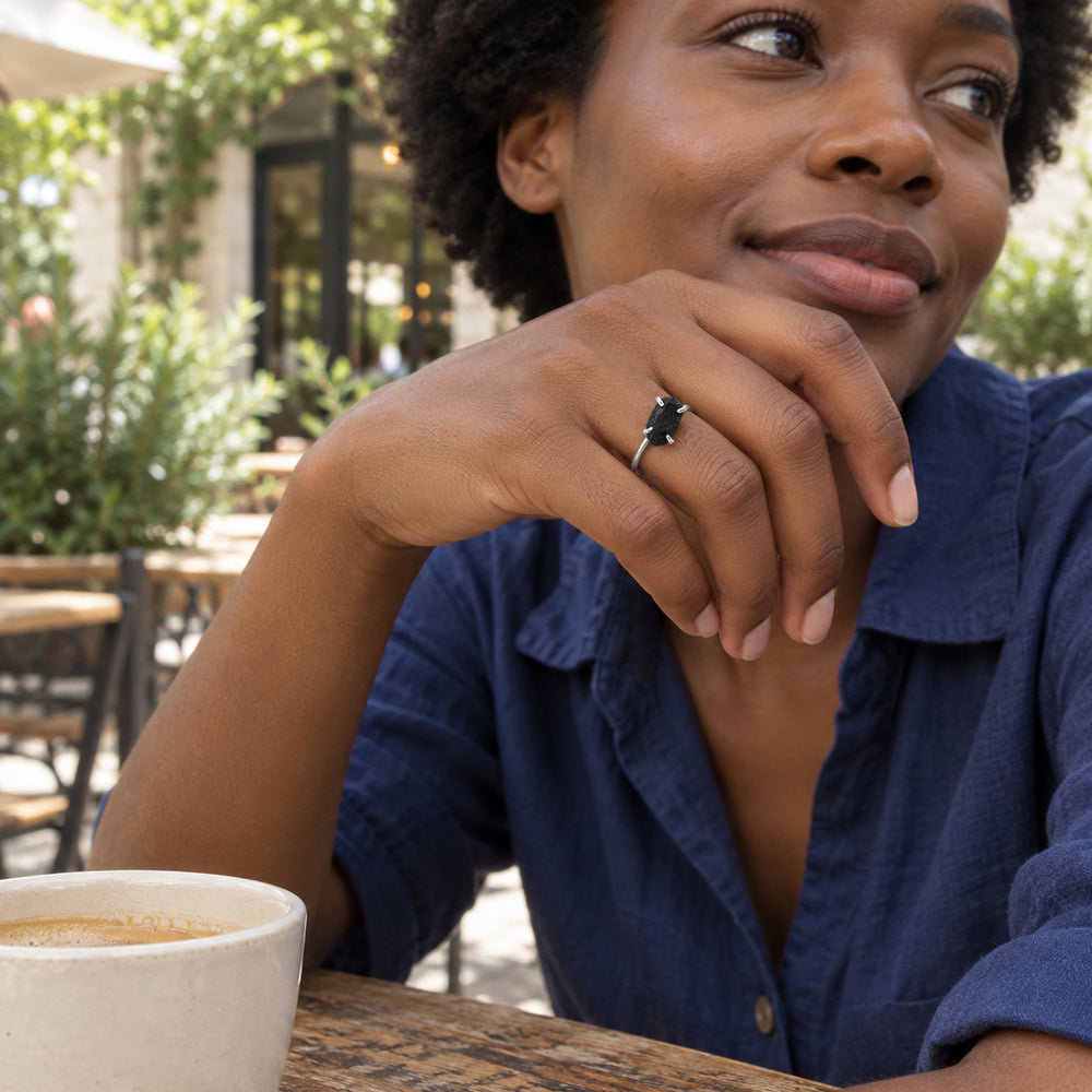 A silver ring with a raw black obsidian stone is worn on a hand at an outdoor cafe beside a cream ceramic cup.