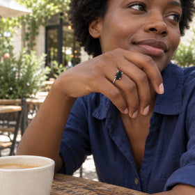 A silver ring with a raw black obsidian stone is worn on a hand at an outdoor cafe beside a cream ceramic cup.