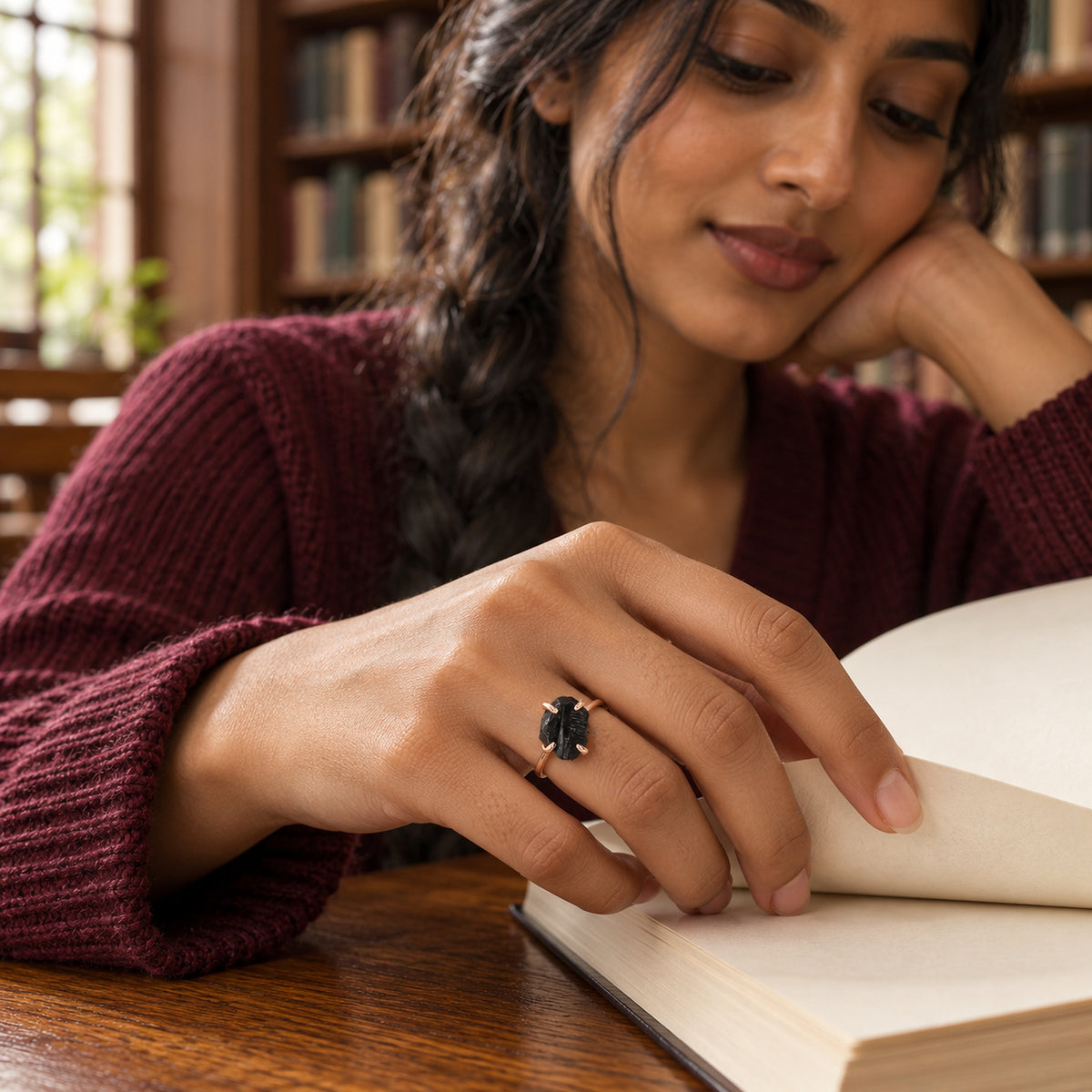 A rose gold vermeil ring with a rough black stone is worn on a hand resting on an open book at a wooden library table.
