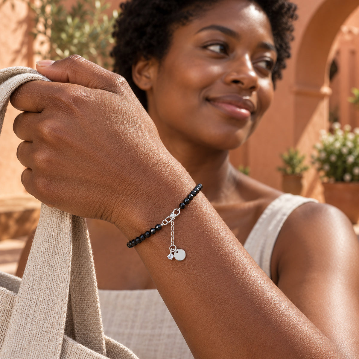 A woman holds a linen bag in a terracotta courtyard while wearing the black bead bracelet with silver fittings.