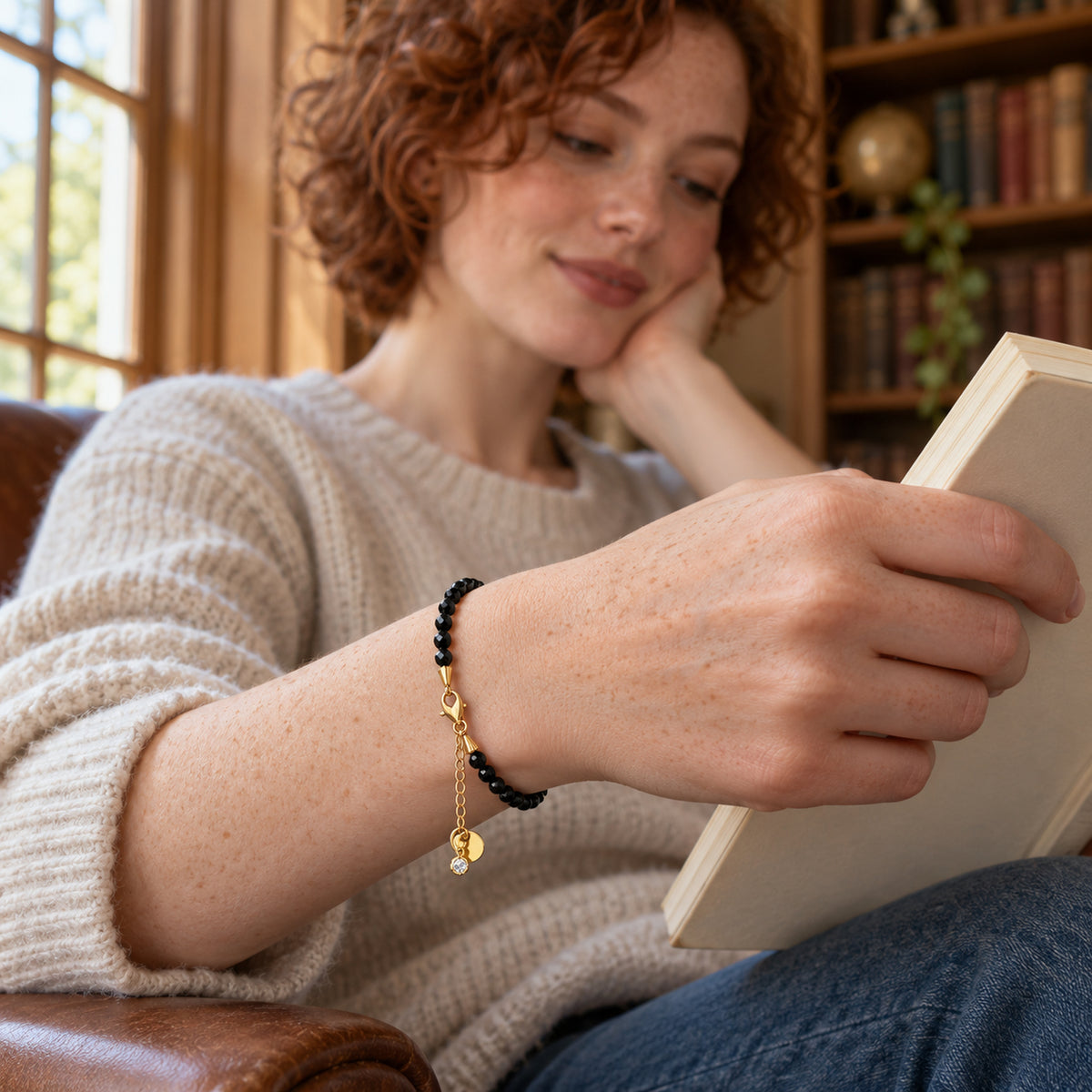 A black obsidian bracelet with yellow gold vermeil hardware circles a freckled wrist beside an open book.
